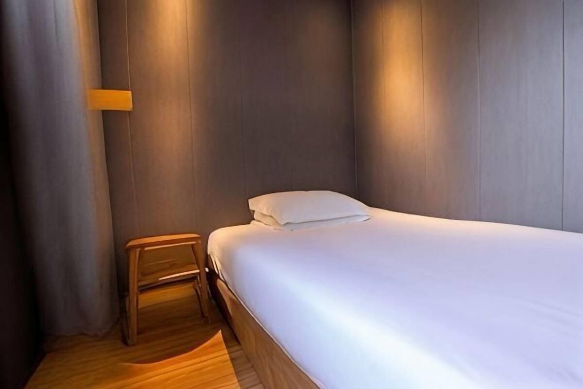 Un lit simple avec un matelas et des oreillers blancs, une table de chevet en bois et un luminaire fixé au mur.