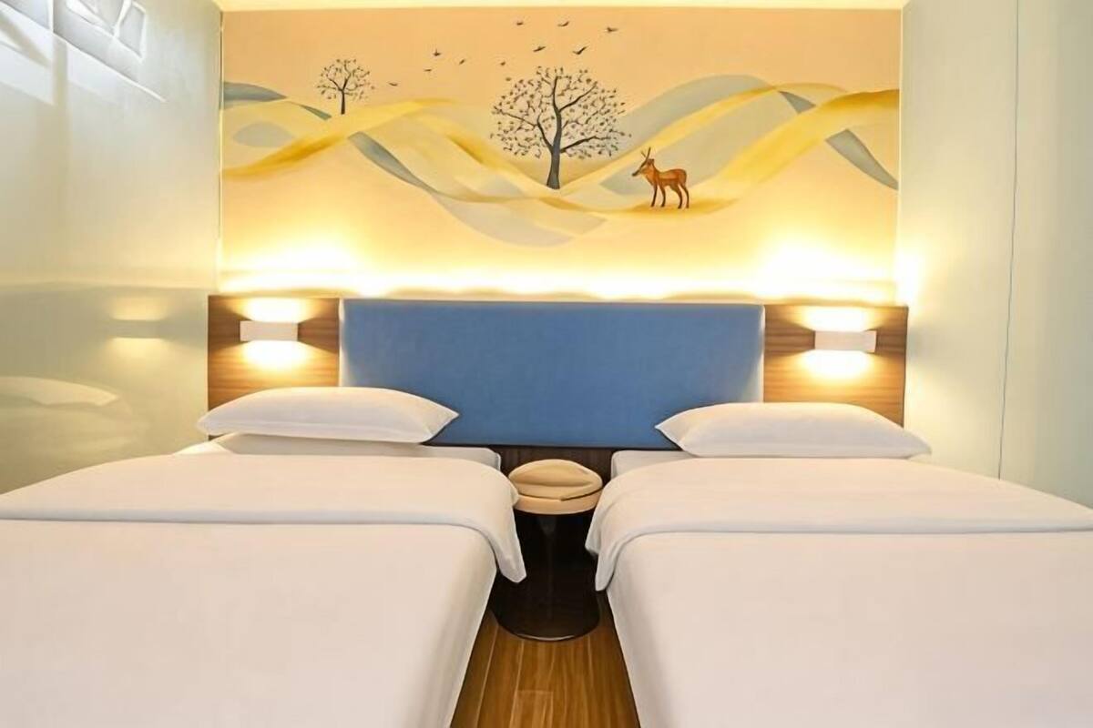 Une chambre d’hôtel avec deux lits, une tête de lit bleue et une fresque murale représentant un arbre et des cerfs.