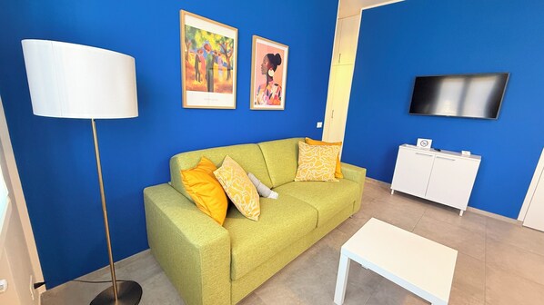 Un salon avec un canapé vert, une table blanche, un lampadaire, un téléviseur et un meuble.