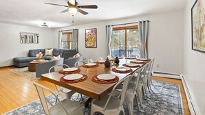 A dining area with a long wooden table set for six, surrounded by white chairs. There is a ceiling fan above, and the room has large windows with curtains. - Near Ithaca!Hot Tub! LRG Home!Sleeps 12! Dogs ok! (Newfield)
