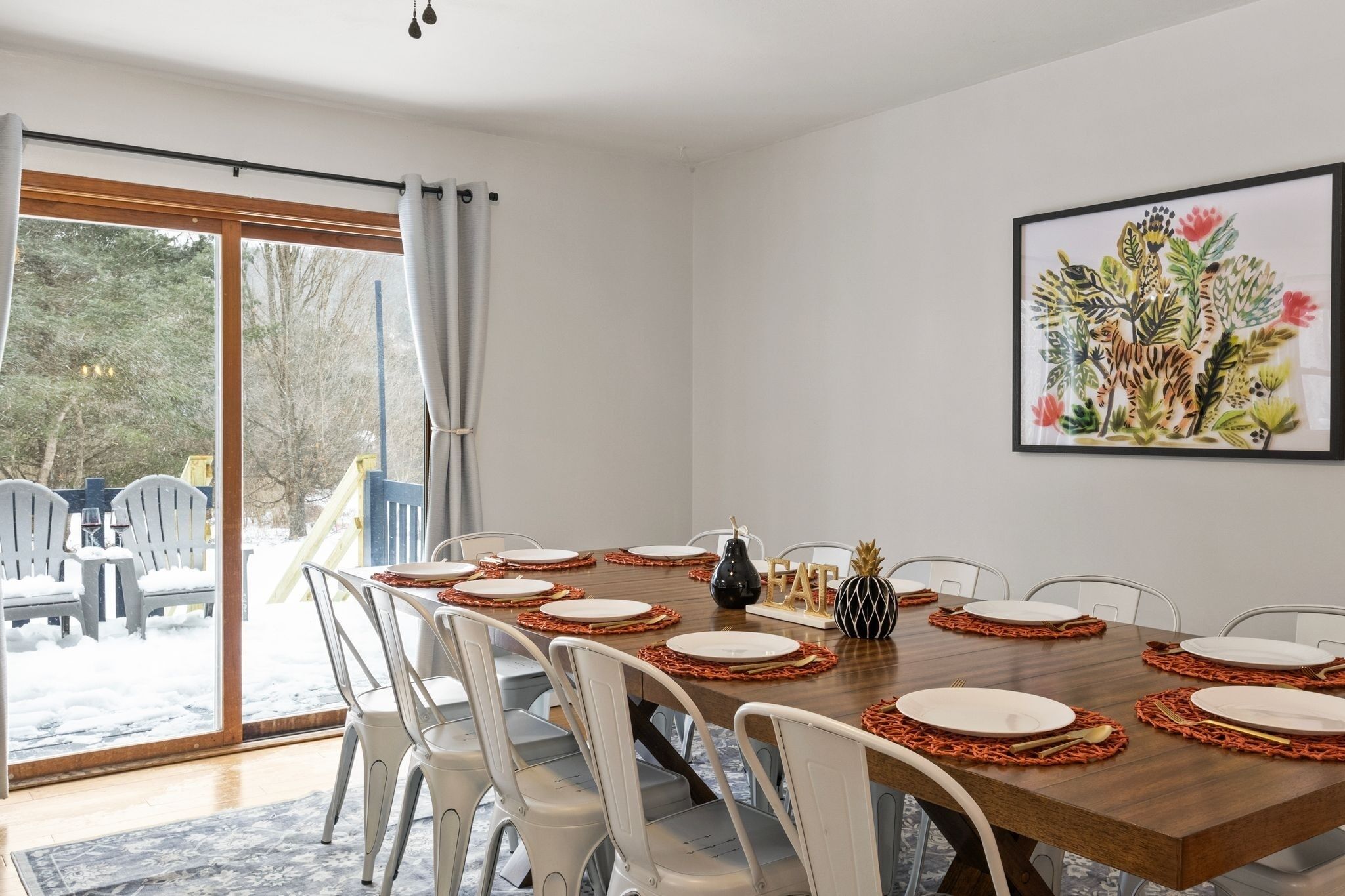 A dining area with a wooden table set for six, white chairs, and a large window with curtains.