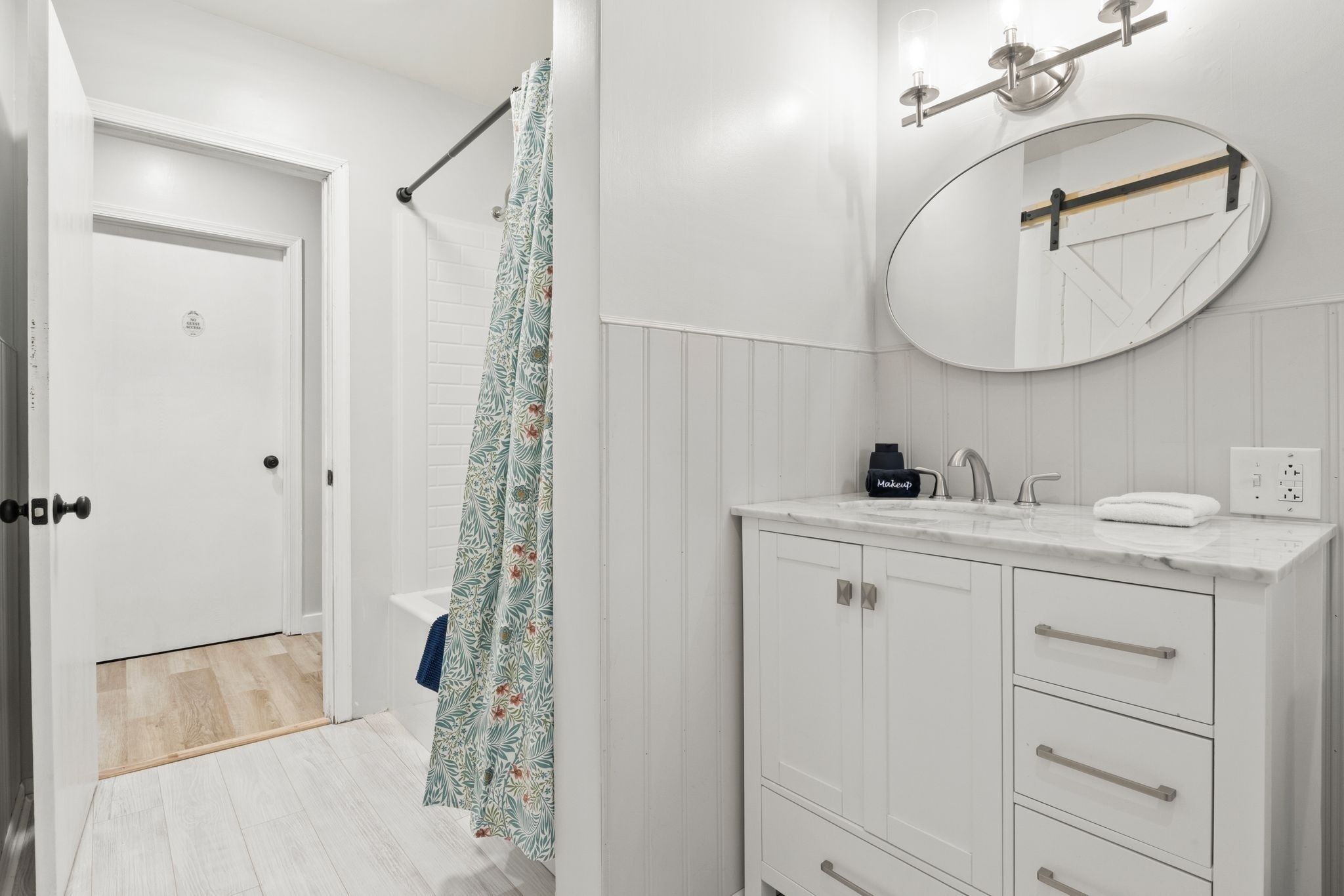 A bathroom with a white door, a shower with a floral curtain, a white vanity with a mirror, and a towel on the counter.
