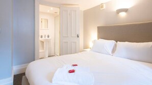 Une chambre d’hôtel avec un grand lit, du linge de lit blanc, une table de chevet, une lampe fixée au mur et une porte donnant sur une salle de bain.