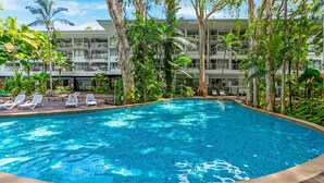 Una piscina con sillas de descanso, rodeada de vegetación tropical y un edificio de varios pisos.