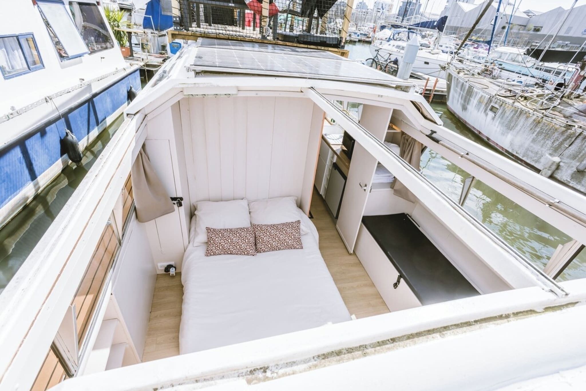 Een compact bootje met een klein bed, een raam en uitzicht op de jachthaven.