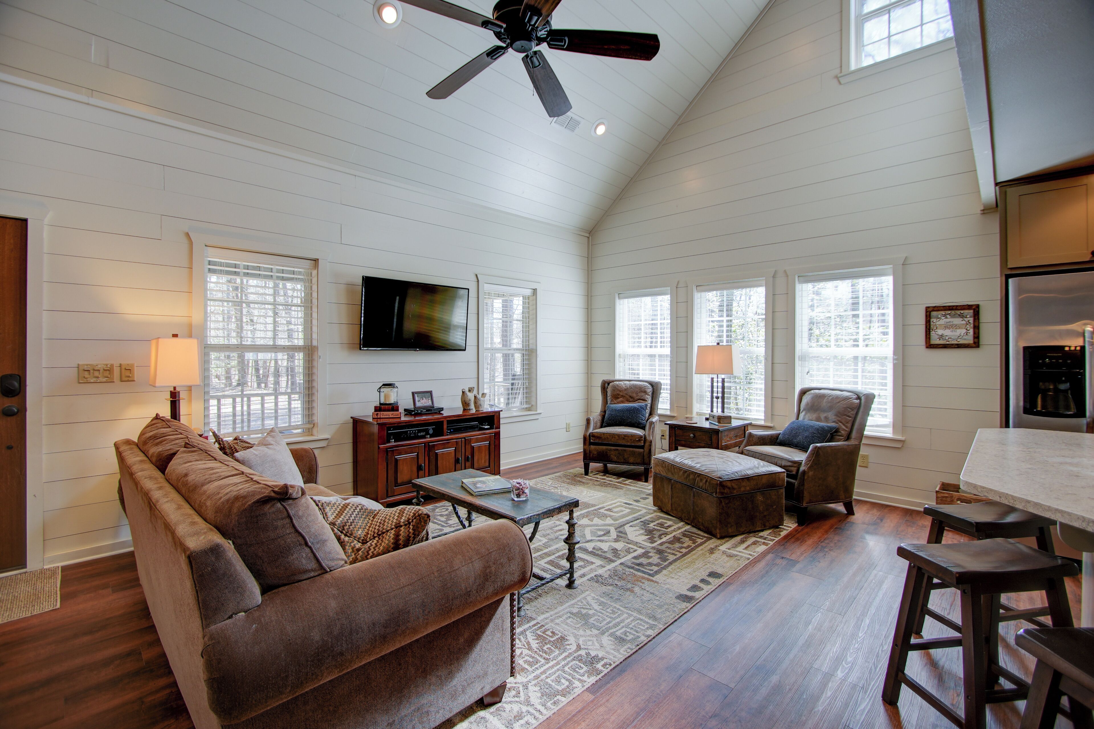 A spacious living room with a high ceiling fan, wooden floors, and a large TV mounted on the wall.