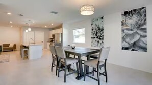 A spacious living area with a dining table, chairs, a kitchen in the background, and a large floral artwork on the wall. - Palm Breeze Lux Stay - Port St. Lucie (Port Saint Lucie)