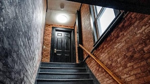 A staircase with a wooden handrail leading to a black door, surrounded by brick walls. - The Cider Loft (Waterbury)