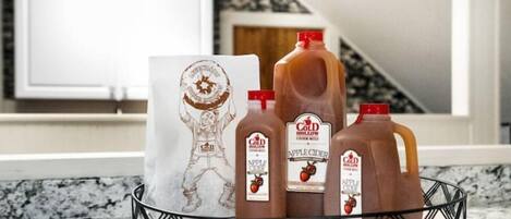 Una encimera de cocina con una cesta metálica que contiene tres botellas de sidra de manzana y una bolsa de papel con un logotipo.