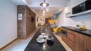 Una cocina moderna con cubierta negra, microondas y cafetera. Se observa una pared de ladrillo decorativa y una zona de estar al fondo.
