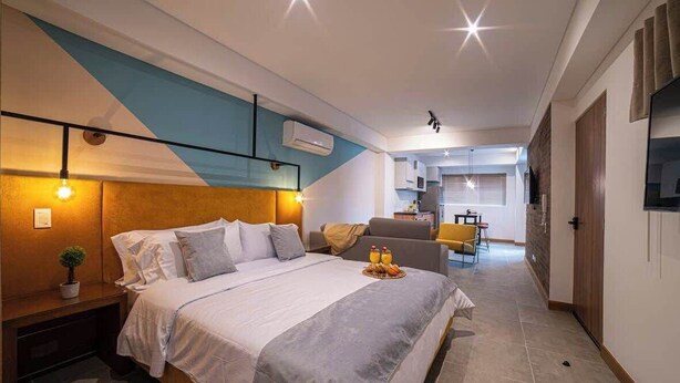 Un dormitorio moderno con cama, mesita de noche y una planta pequeña. Se observa una cocina al fondo con una mesa de comedor y sillas.