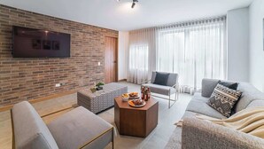 A modern living room with a brick wall, a flat-screen TV, a coffee table with food, and a grey sofa. - E201 - Spacious 2-bed / 2-bath, A/C & Fast WiFi (Cali)