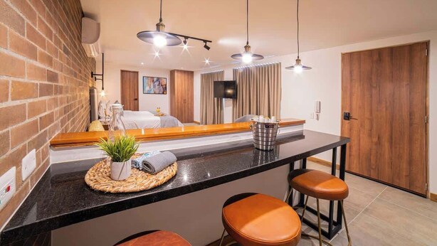 A modern kitchen with a black countertop, two brown bar stools, a hanging light fixture, and a brick wall. - E103 - Brand-New Quiet & Comfortable Studio Unit (Cali)