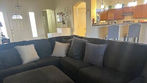 A dark-colored sectional sofa with striped and solid pillows, a small ottoman, and a kitchen area in the background with wooden cabinets and a countertop. - 2-bedroom clean home in Sierra Vista, WiFi, AC, covered patio & shaded backyard. (Sierra Vista)