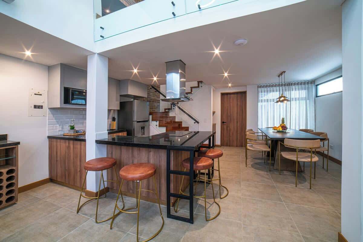 Una cocina moderna con una isla central, taburetes de bar, una zona de comedor con mesa y sillas, y una escalera que sube a un nivel superior.