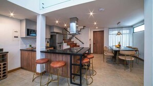 Eine moderne Küche mit einer zentralen Kochinsel, Barhocker, einem Essbereich mit Tisch und Stühlen sowie einer Treppe, die zu einer oberen Ebene führt.