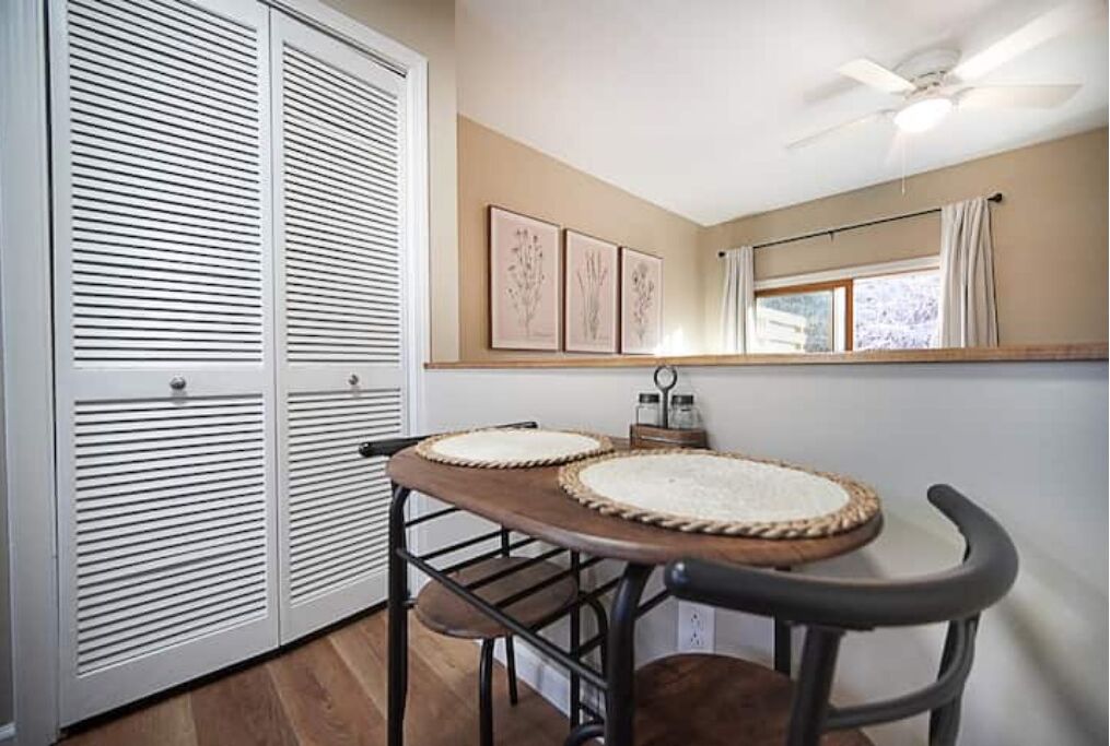 A room with a wooden table, two chairs, a ceiling fan, a window with curtains, and a set of white louvered doors.