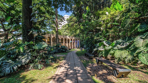 A pathway leading to a building with arched windows, surrounded by lush greenery and a bench.