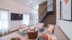 A modern living room with a sofa, two armchairs, a coffee table, and a staircase against a brick wall.