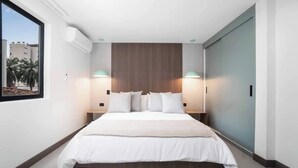 A modern bedroom with a large bed, two bedside lamps, a wooden headboard, and a window with a view of palm trees. - Hygge 301 - 2-Bed Beauty | Balcony | Near Granada (Cali)