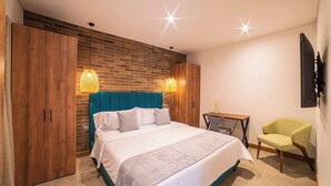 A modern bedroom with a brick accent wall, a teal upholstered headboard, a large bed with white bedding, two wall-mounted lamps, a small side table, and a chair. - Luxury 3-Bed | Walk to Boulevar & Granada | E101 (Cali)