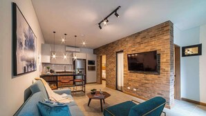 A modern living room with a sofa, armchair, and coffee table, connected to a kitchen with a brick accent wall and a TV mounted above it. - Luxury 3-Bed | Walk to Boulevar & Granada | E101 (Cali)