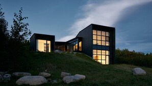 Ein modernes Haus mit großen Fenstern und dunkler Fassade, gelegen auf einem grasbewachsenen Hügel mit Steinen.
