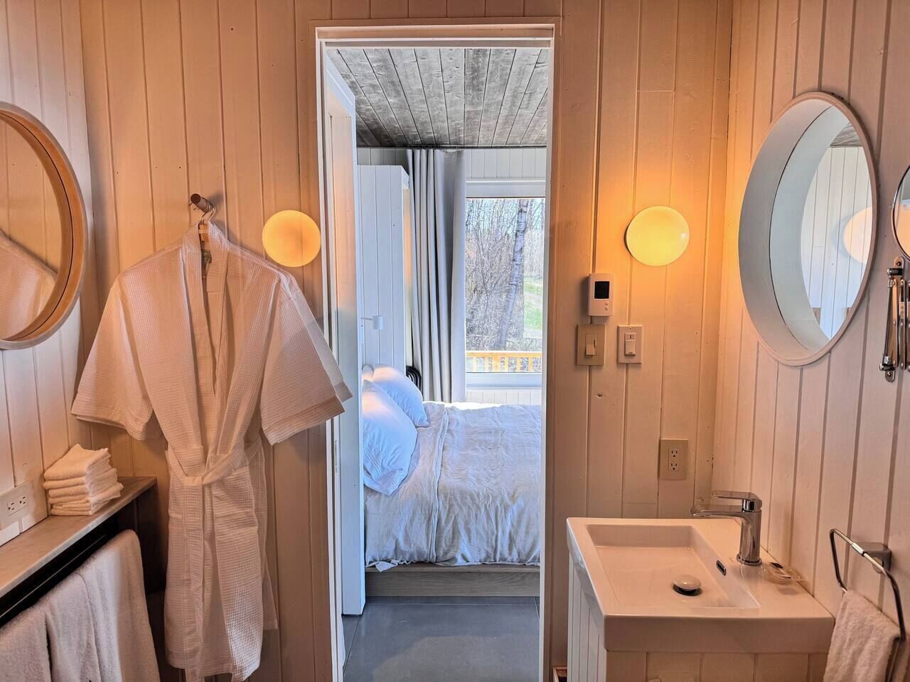 A hotel room with a bed, a bathroom with a sink, and a mirror. There is a robe hanging on the wall and a towel on the floor.