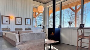Una sala de estar moderna con sofá, mesa de centro, chimenea y un amplio ventanal con vistas a árboles.