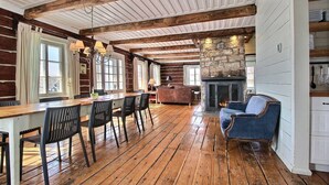 A spacious room with a wooden floor, a stone fireplace, a dining area with a long table and chairs, and a blue armchair.