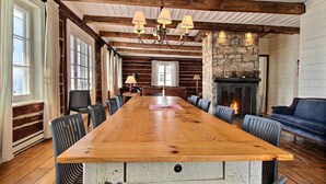 A large wooden dining table with chairs, a stone fireplace, and a blue sofa in a rustic room with exposed wooden beams and large windows.