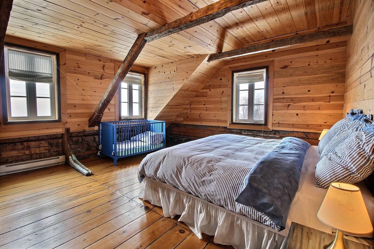 Une chambre dans une cabane en bois, avec un plafond en pente, un lit, un berceau bleu et une fenêtre.