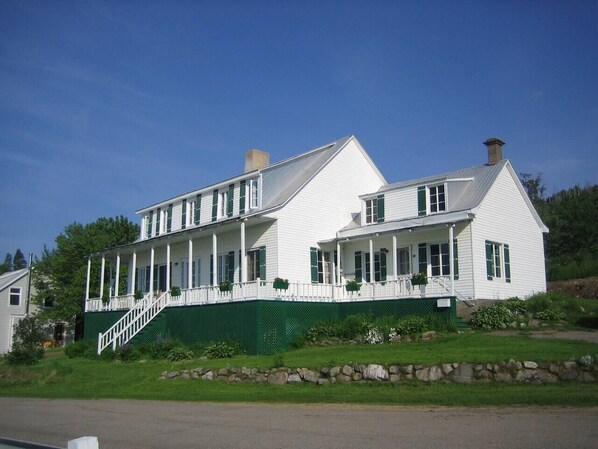 A two-story white house with green shutters, a front porch, and a stone retaining wall.
