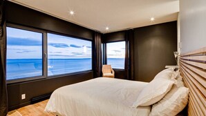 A bedroom with a large bed, a view of the sea through two windows, a chair, and a nightstand. - Chalet Marée Basse | Charlevoix (La Malbaie)