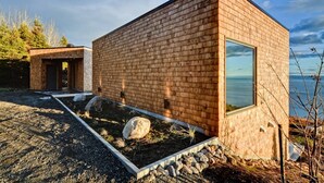 A modern wooden cabin with a large window, a stone pathway, and a view of the water. - Chalet Marée Basse | Charlevoix (La Malbaie)