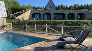 Zona de piscina con sillones de descanso, un edificio de piedra con ventanas de arco y una terraza de madera.