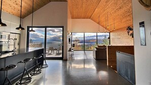 A modern kitchen with a bar area, large windows offering scenic views, and a high ceiling with wooden beams.