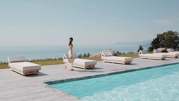 A poolside area with lounge chairs and a mattress on a wooden deck, overlooking a body of water and distant mountains.
