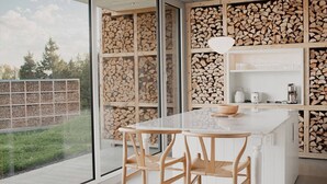 A modern kitchen with a large island, wooden chairs, and a view of a backyard with stacked firewood.