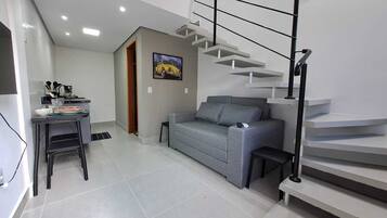 A modern living space with a grey sofa, a dining area with a table and chairs, and a staircase with a metal railing.