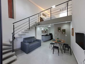 A modern living space with a staircase, grey sofa, dining table with chairs, kitchen area, and a flat-screen TV. - LOFTS INDAIATUBA (Indaiatuba)