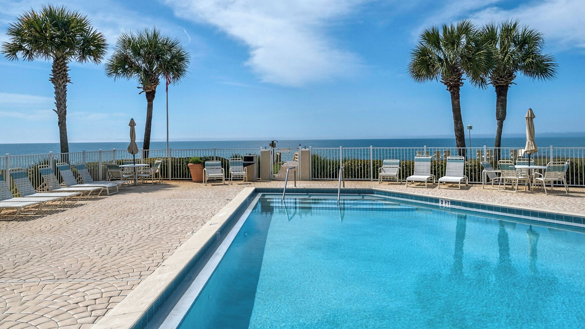 A pool area with lounge chairs and tables, a fenced pool, and palm trees.