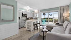 A modern kitchen with white cabinets, stainless steel appliances, and a central island. There is a dining area with a white table and two bar stools. A grey sofa and a small round table are in the living area. - Sand Cliffs 108: Steps to Beach & Pool Access! (Panama City Beach)