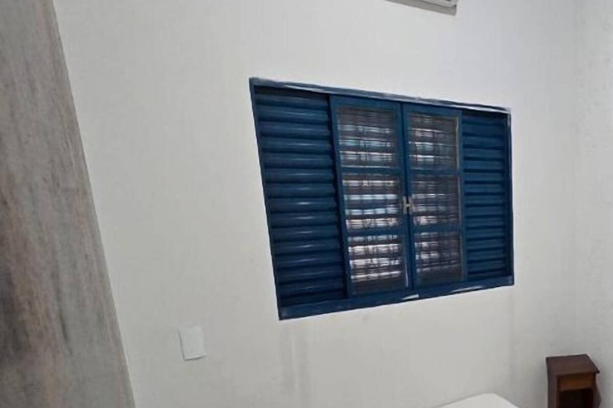 A small, simple room with a single bed, a window with blue shutters, and a small table.