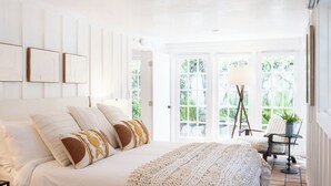 A bedroom with a large bed, a chair, and a table by a window with a view of greenery. - Buttterfly Beach Dream Cottage (Montecito)