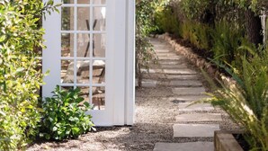 A pathway leading to a white structure with a glass door, surrounded by greenery and trees. - Buttterfly Beach Dream Cottage (Montecito)