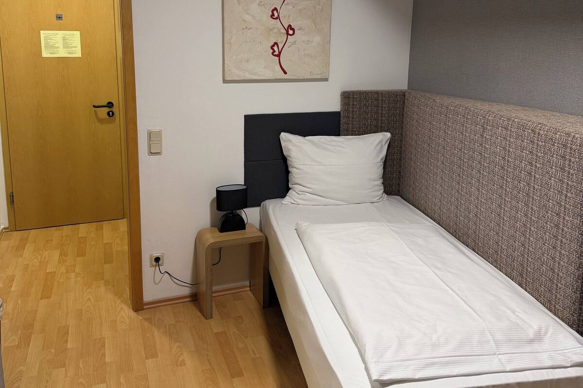 A hotel room with a wooden floor, a bed with white bedding, a nightstand with a lamp, a headboard, and a wall-mounted artwork.