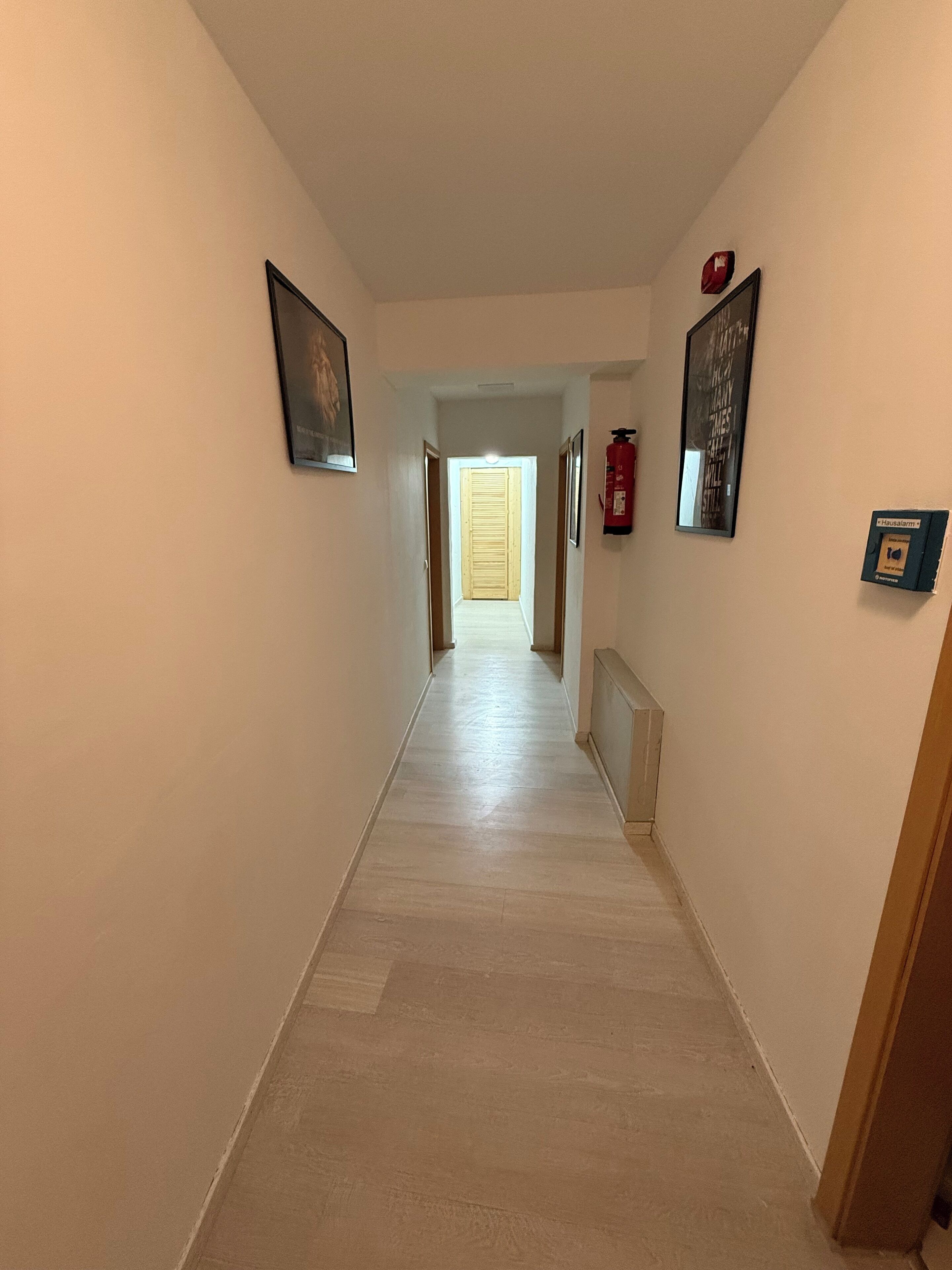 A hallway with a wooden floor, framed pictures on the walls, a fire extinguisher, and a door at the end.
