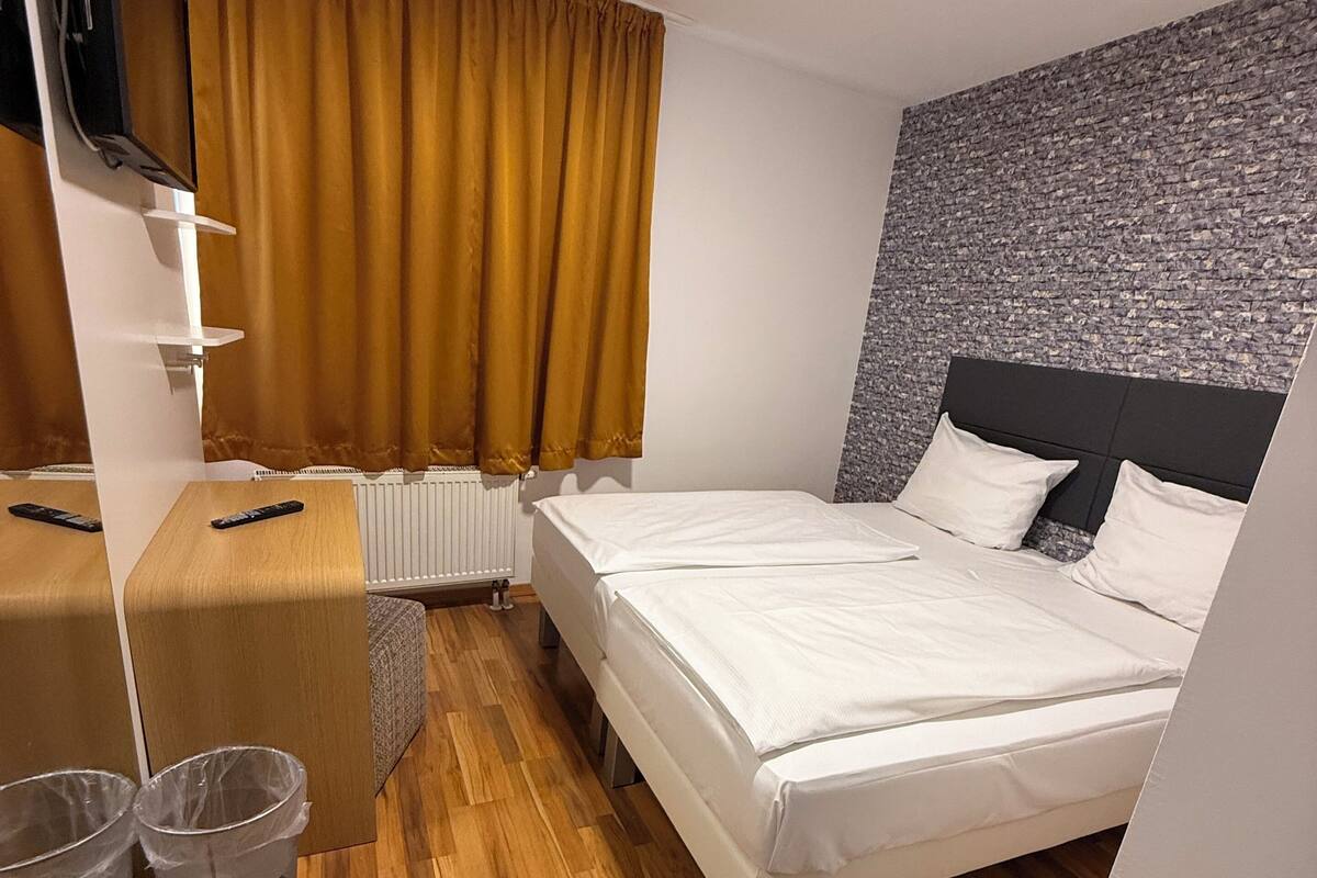 A hotel room with a wooden floor, a bed with white linens, a television mounted on the wall, and a window with yellow curtains.