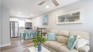 Una sala de estar moderna con un sofá beige, una mesa de centro con una planta, y una cocina al fondo.